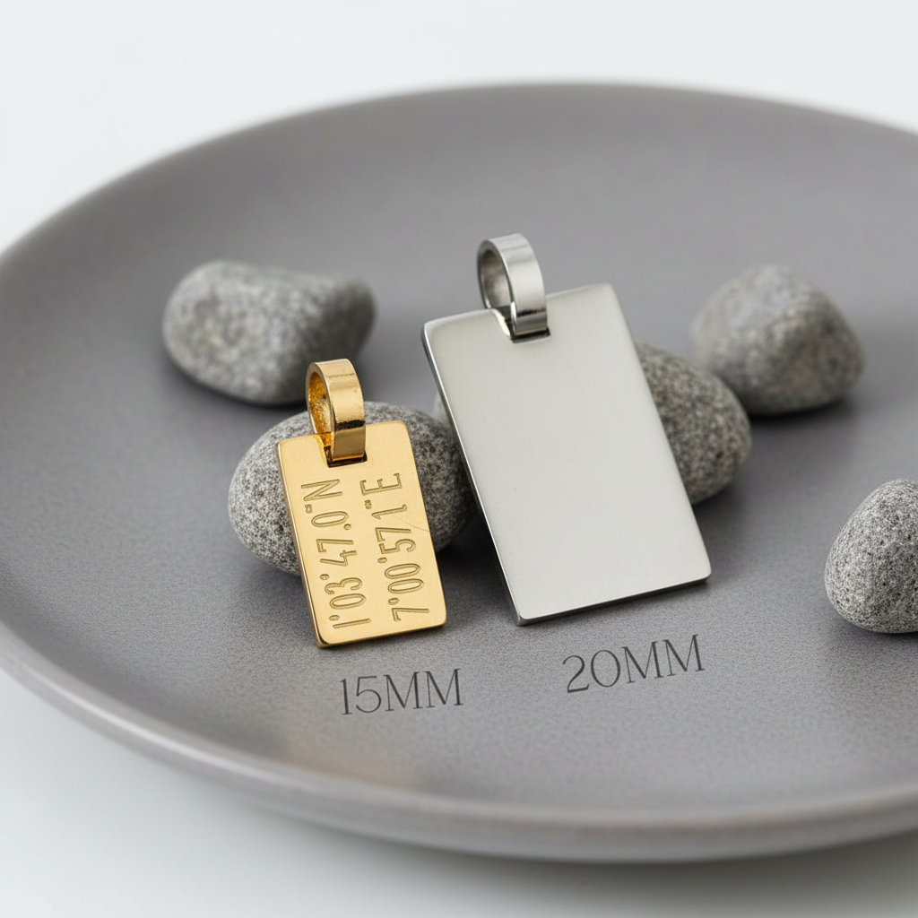 Zwei rechteckige Schmuckanhänger aus Edelstahl auf grauem Teller mit Dekosteinen, links ein goldfarbener Anhänger mit Koordinaten-Gravur in 15 mm, rechts ein silberfarbener Anhänger in 20 mm ohne Gravur, minimalistische Produktdarstellung.